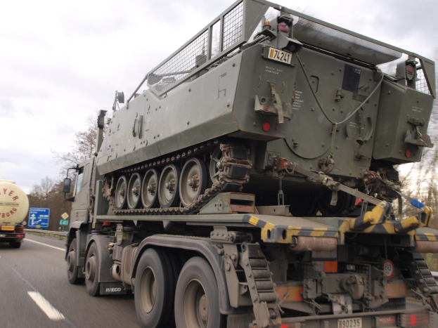 Militärlastwagen transportiert einen Panzer auf seiner Ladefläche, fährt auf einer Autobahn mit Bäumen, hölzernen Barrieren und einem klaren blauen Himmel im Hintergrund.