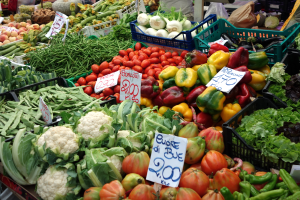 Ein belebter Bauernmarkt mit verschiedenen Gemüsen wie Tomaten, Paprika, Blumenkohl und grünen Bohnen in Körben, Preisetiketten sichtbar und einige Menschen anwesend.