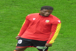 Ein Mann in rotem T-Shirt und schwarzen Shorts steht auf einem Fußballfeld, lächelt und ist bereit, als Flügelstürmer zu spielen.