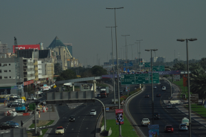 Luftaufnahme einer Stadt mit Gebäuden, Türmen, Kraftfahrzeugen, Werbetafeln, Schildern, Straßen, Bäumen, Straßenlaternen, Menschen, Absperrgittern und Informationsschildern unter einem klaren Himmel.