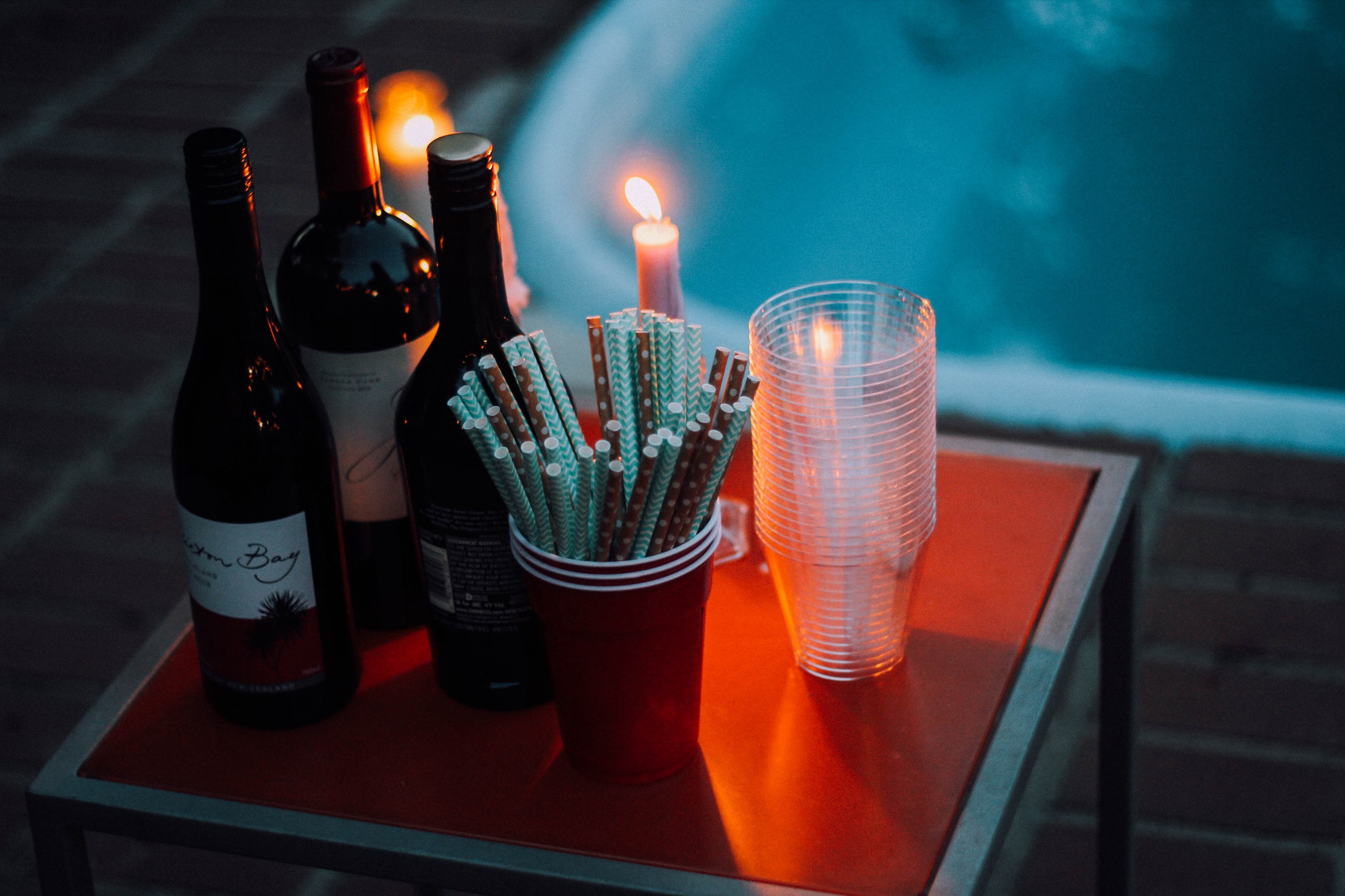 Zwei Flaschen Wein, Gläser mit Strohhalmen und eine brennende Kerze auf einem Tisch neben einem Swimmingpool.
