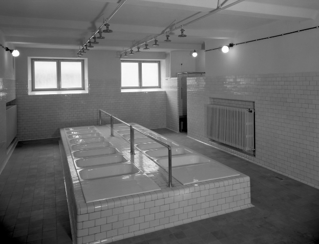 Schwarzes und weißes Foto einer öffentlichen Toilette mit einer Reihe von Waschbecken, Fliesenwänden, Fenstern, Lampen und Rohren.