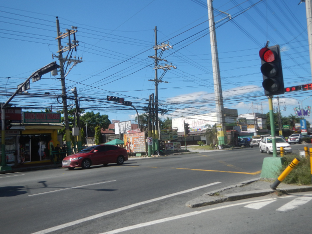 Ein rotes Auto fährt auf einer Straße neben einer Ampel, mit Strommasten und Drähten an den Seiten, Gebäuden, Bäumen und einem klaren blauen Himmel im Hintergrund.