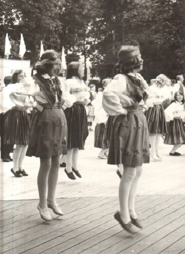 Schwarzes und weißes Foto von Frauen in traditioneller bayrischer Tracht, die auf einem Holzfußboden im Freien tanzen, umgeben von Bäumen und Fahnen.