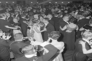 Ein Schwarz-Weiß-Foto einer Gruppe von Menschen, die auf einer Tanzfläche tanzen, einige halten Kameras in der Hand, mit Treppen und Lichtern im Hintergrund.