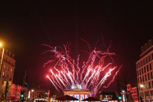 Eine belebte Stadtstraße in Berlin an Silvester, voller Menschen, Fahrzeuge und Gebäude, erhellt von Feuerwerk und Gebäudebeleuchtung, die eine festliche Stimmung schaffen.